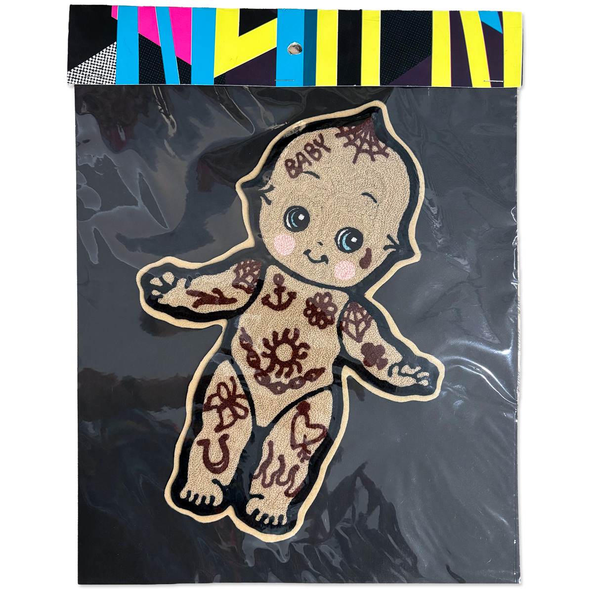 A decorative embroidered patch shaped like a baby figure, shown inside clear plastic packaging with a black backing and a colorful striped header card. The patch has a tan base with dark brown tattoo-style patterns and the word 'BABY' embroidered on the forehead.