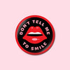 Button with the text "Don't tell me to smile" surrounding an illustration of lips.
