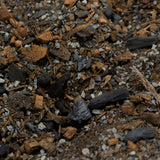 A close-up of a soilless potting mix blend containing dark charcoal, brown bark chips, and small white gravel, with a metal scoop visible on the left edge.