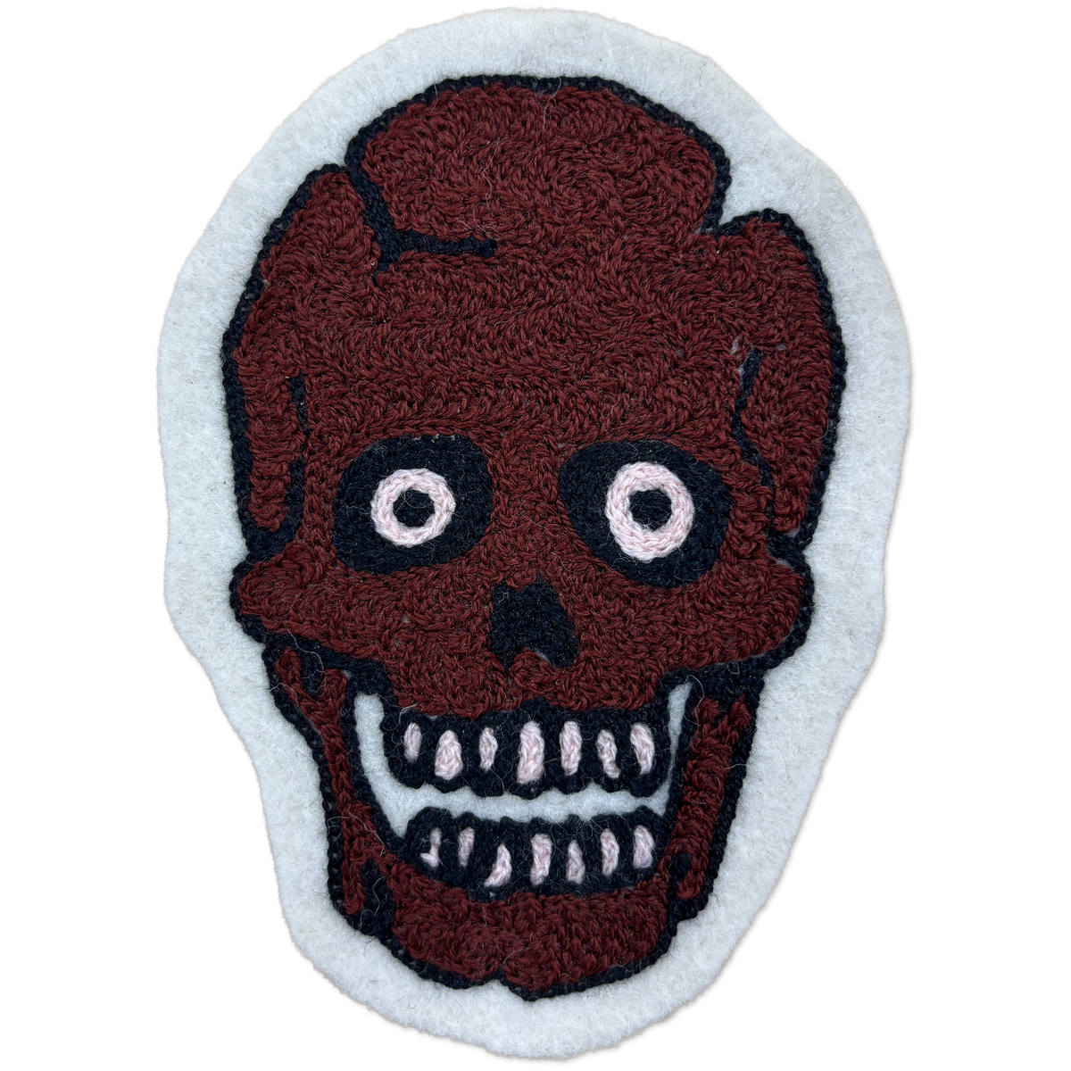 A handcrafted chainstitch patch featuring a stylized skull with a dark reddish-brown textured face, black outlines, and white details, set against a light gray background.