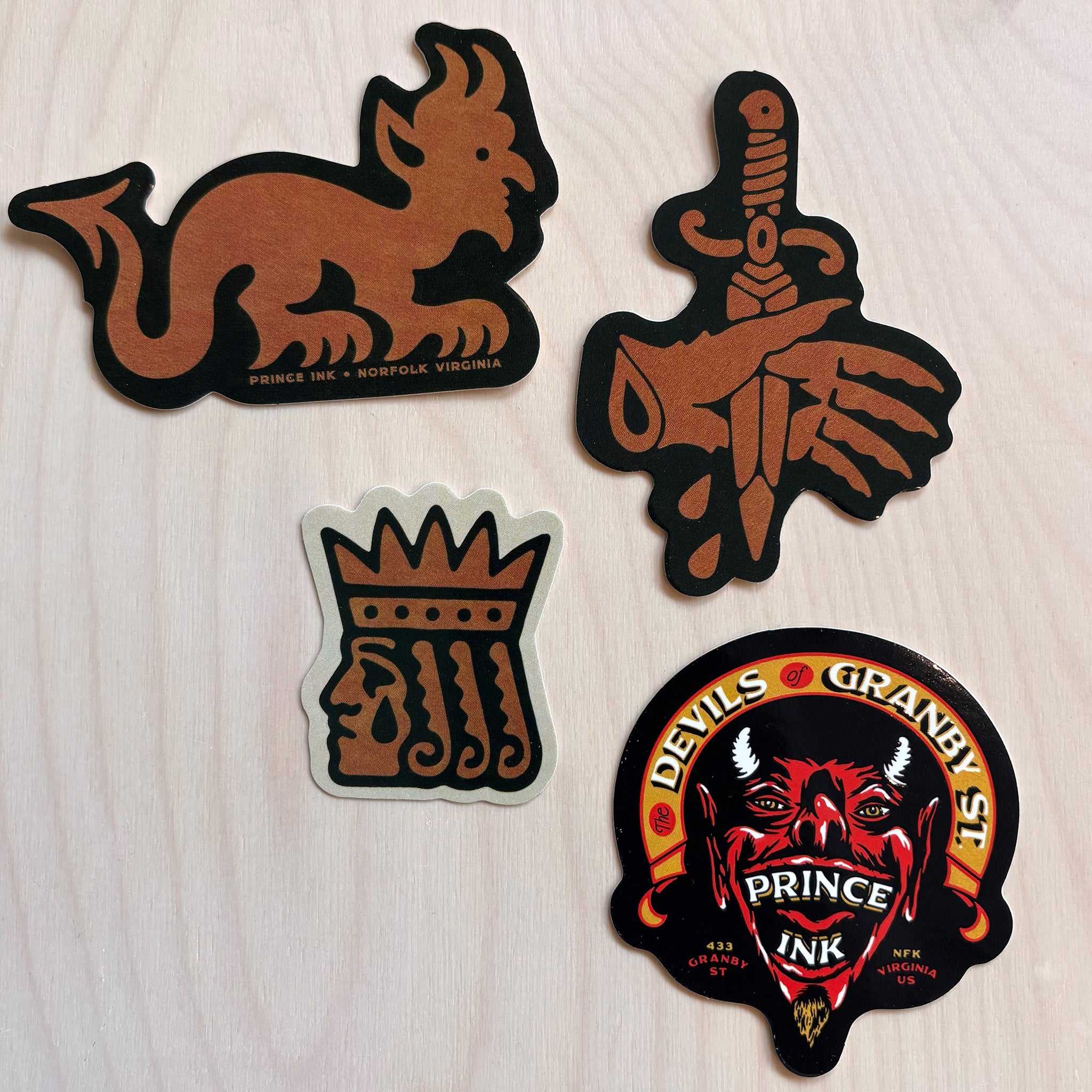 Four die-cut stickers displayed on a light wood background. The set includes a brown goblin sticker, a brown hand holding a sword, a brown crown , and a red and black devil face sticker with 'Devils of Granby St. - Prince Ink'.